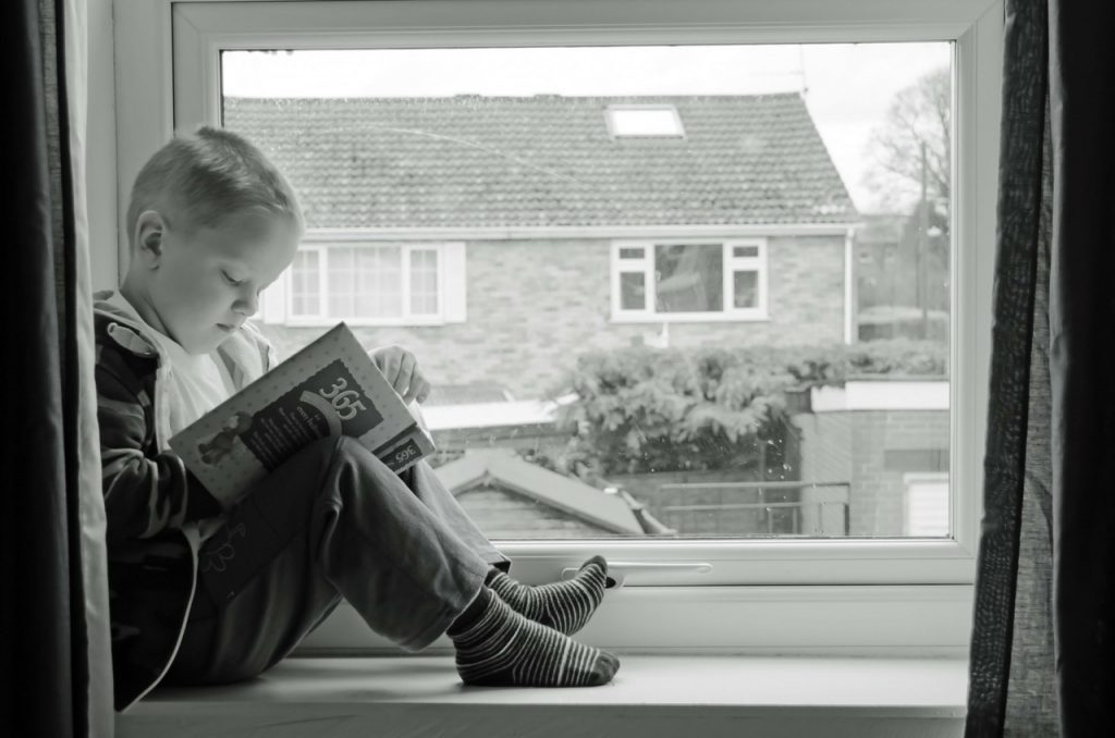 A kid reading story book sitting on a window sill
